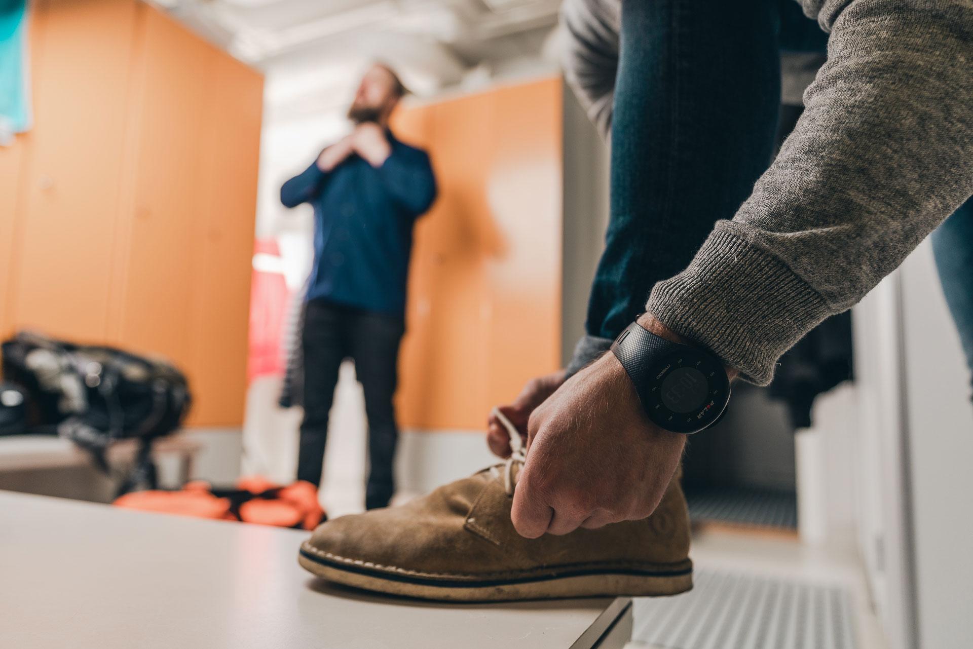 A man ties shoes in the locker room and another man in the background corrects his collar.