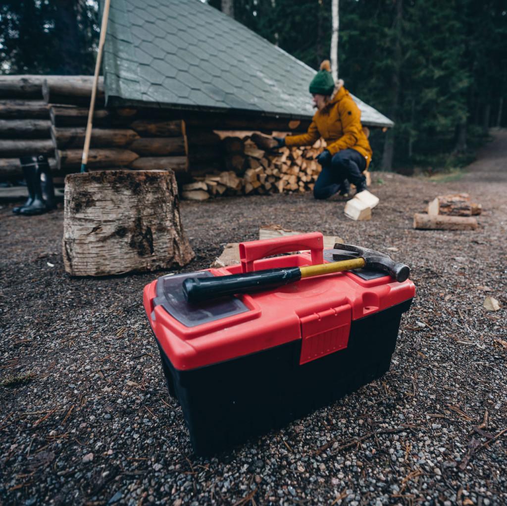 In the foreground a toolbox with a hammer on it, and in the background a woman in front of a wooden shed.