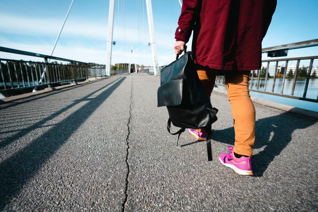 Kuvassa näkyy naisen alavartalo takaapäin, kun tämä kävelee sillalla laukku kädessään.