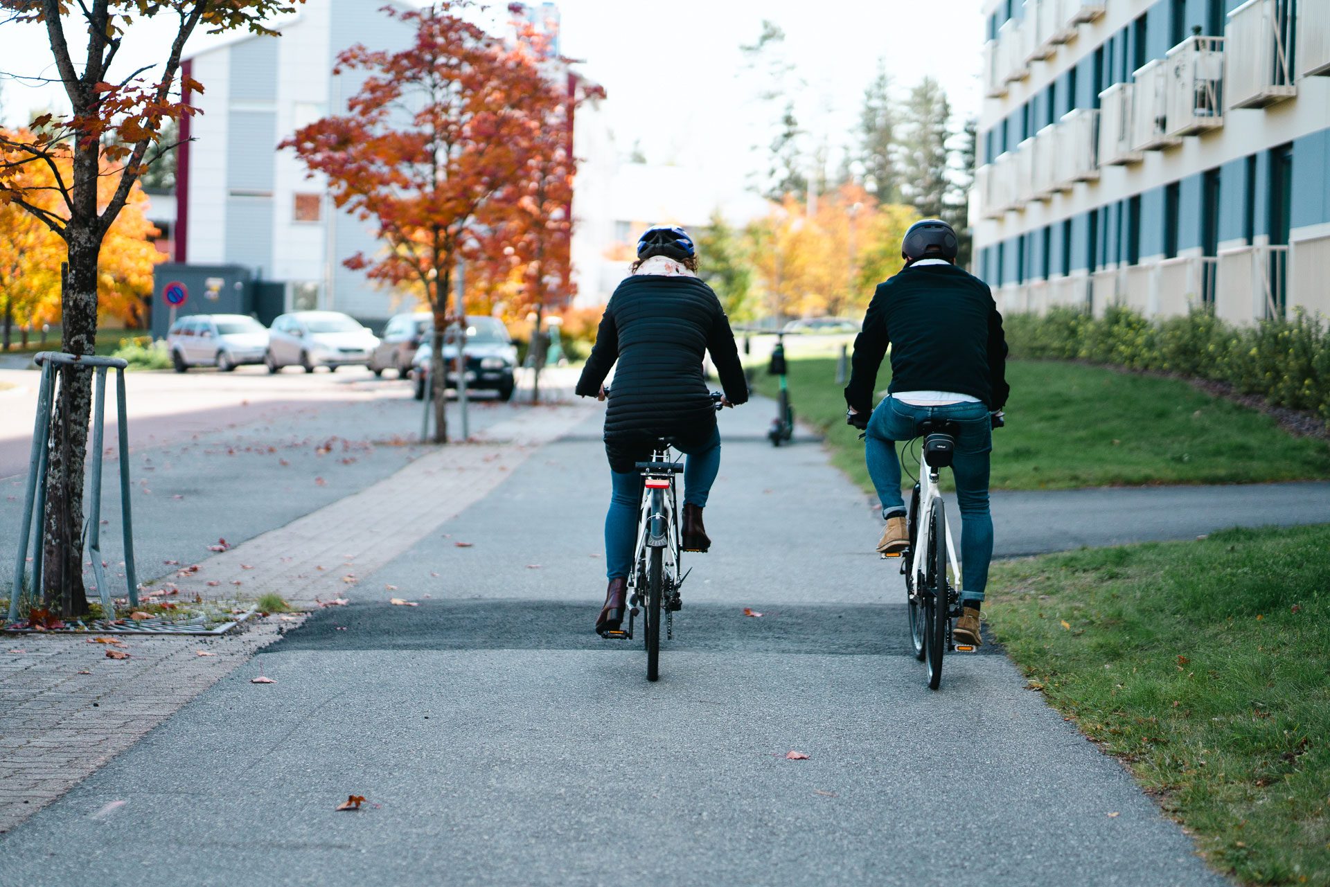 Kuvassa kaksi henkilöä ajaa polkupyörillä pois päin kamerasta kaupunkimaisemassa.