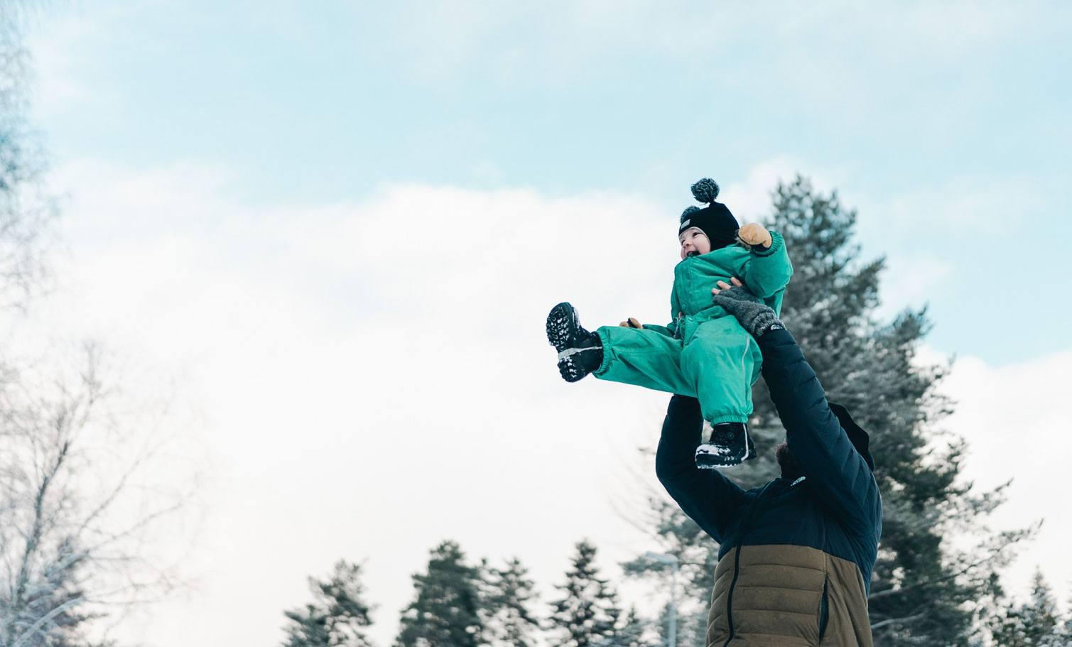 Kuvassa mies nostaa talvihaalariin puettua lasta ylös kohti sinistä taivasta.