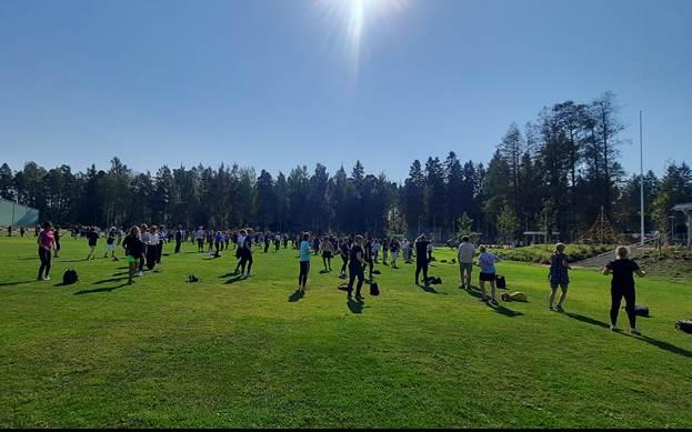 Kuvassa on suuri joukko ihmisiä jumppaamassa aurinkoisella nurmikentällä.