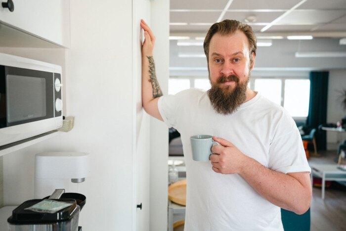 Mies venyttelee kättään jääkaapin oveen nojaten kahvikuppi toisessa kädessä.