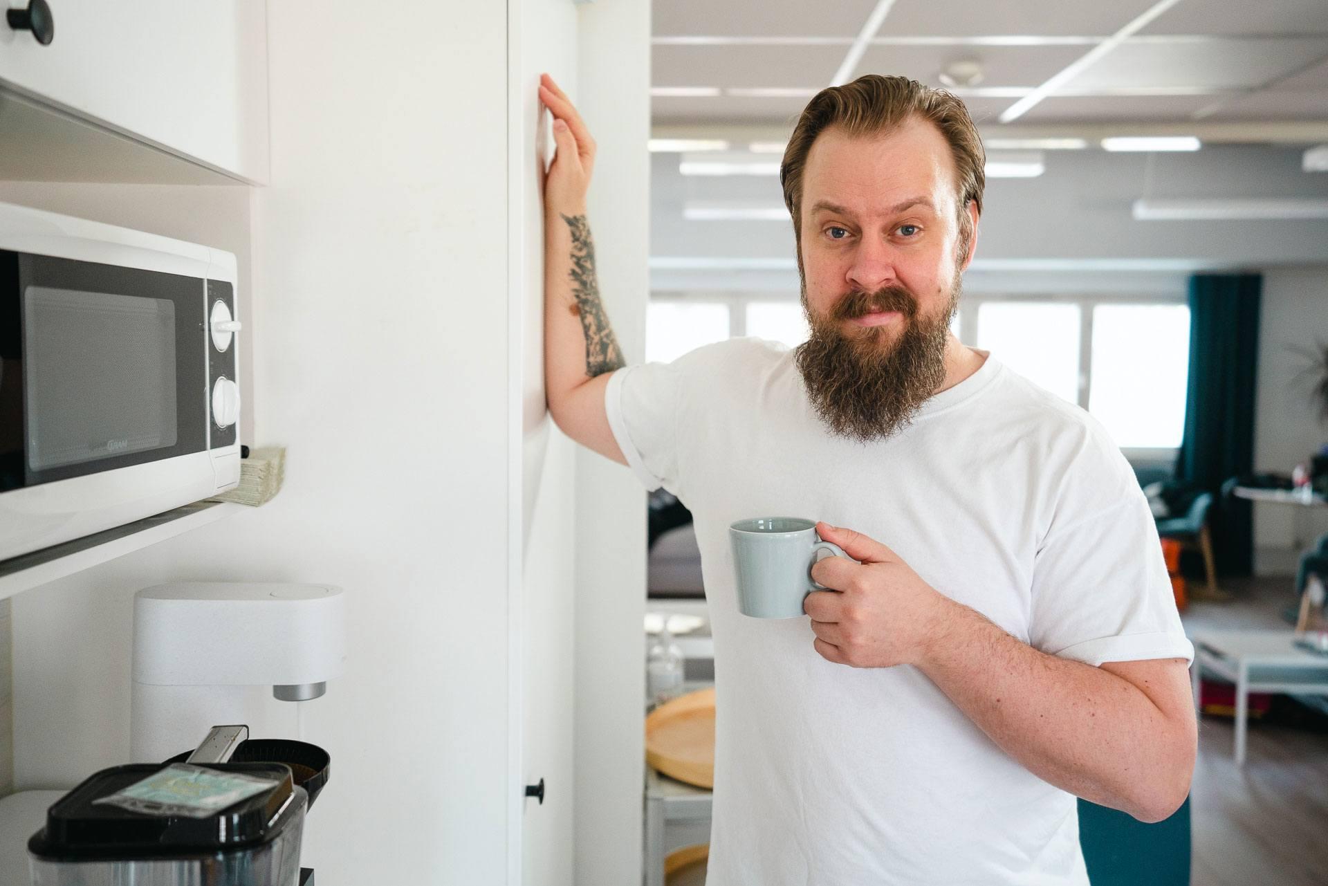 Mies venyttelee kättään jääkaapin oveen nojaten kahvikuppi toisessa kädessä.