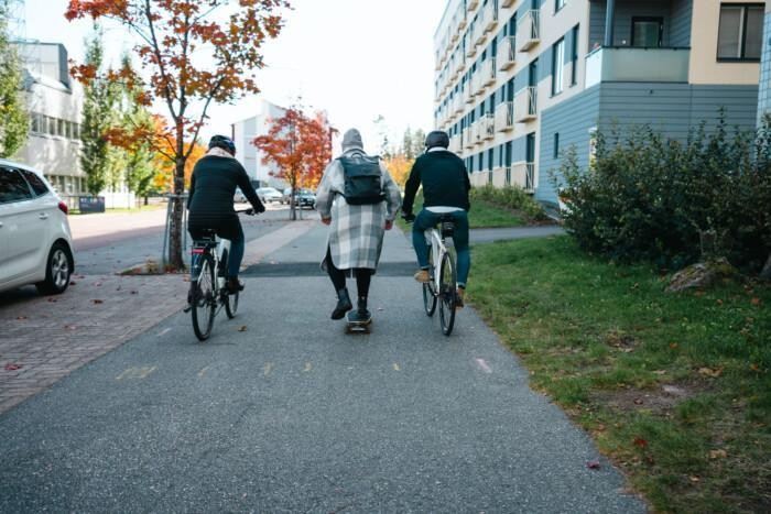 Kuvassa takaapäin kuvattuna kaksi pyöräilijää, joiden välissä yksi henkilö liikkuu skeittilaudalla. Taustalla näkyy kaupunkimaista ympäristöä.
