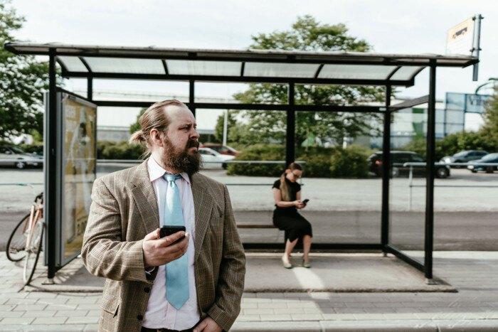Kuvassa etualalla mies seisoo bussipysäkillä matkapuhelin kädessään. Taustalla nainen istuu pysäkin katoksen alla katsoen matkapuhelintaan.