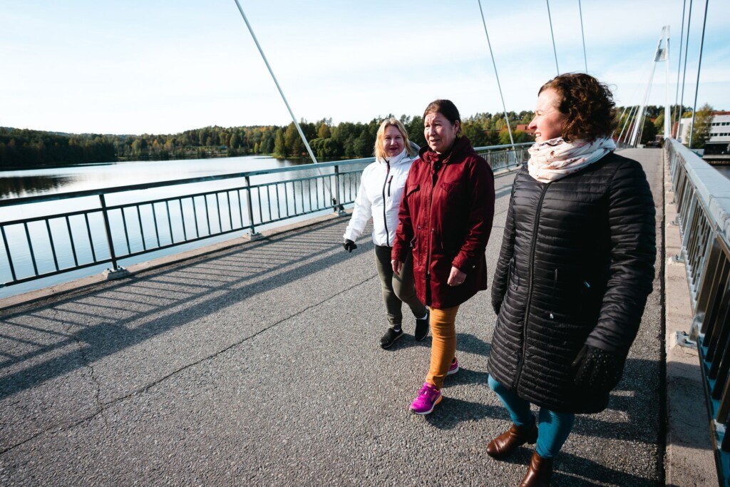 Kolme naista kävelee sillalla. Taustalla näkyy vettä ja puustoa.